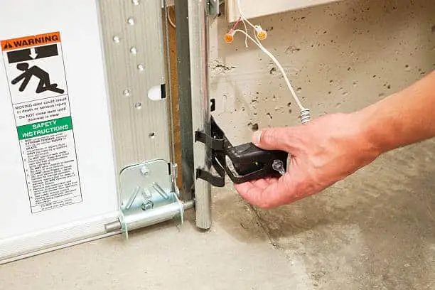 A hand adjusting a garage door safety reversing sensor, with an illuminated green LED to confirm the sensor is lined up with the opposite side sensor and operational. These sensors, required in all new installations, reverse the door when an infrared beam is broken by a child, animal or object. A warning and safety instructions sticker is to the left but slightly soft. There is some paint overspray visible on the sensor and track.