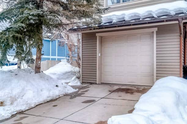 White garage door entrance of home with snow covered roof and yard in winter. Exterior of a house with gray wall siding, wet driveway, and evergreen tree.