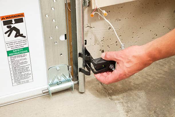 A hand adjusting a garage door safety reversing sensor, with an illuminated green LED to confirm the sensor is lined up with the opposite side sensor and operational. These sensors, required in all new installations, reverse the door when an infrared beam is broken by a child, animal or object. A warning and safety instructions sticker is to the left but slightly soft. There is some paint overspray visible on the sensor and track. Garage Door Repair Eaton Rapids