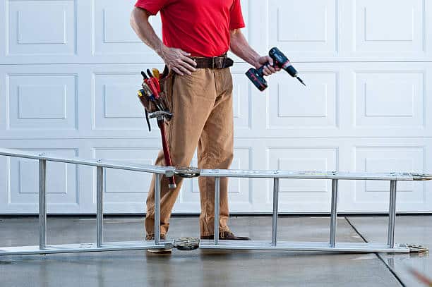 Male handyman with tool belt and ladder working outside. The background is a metal sectional garage door