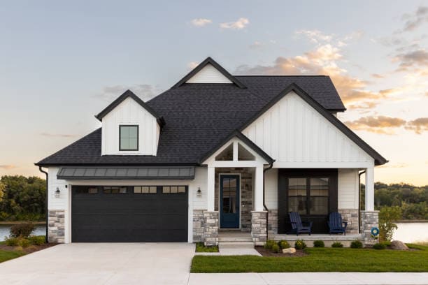 A black and white luxury home with a two car garage and black door, professional landscaping, and a lake in the background at sunset.