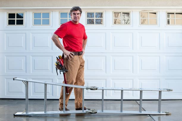 Male handyman with tool belt and ladder working outside. The background is a metal sectional garage door